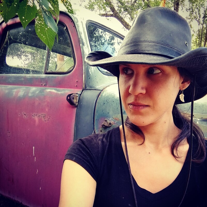 Photo shows Althaea Sebastiani, an olive skinned woman in their late 30's wearing a black v-neck t shirt and a wide brimmed oiled cotton hat. Behind them is a pickup truck from the 1940's, it's door and hood mismatched and creating a colorful background. Tree leaves hang in the corner of the frame.